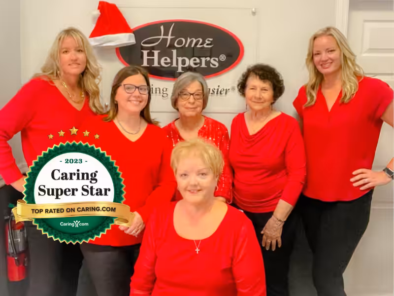 Five women in red tops pose together at a Home Helpers location. A “2023 Caring Super Star” badge appears in the lower left corner. One woman wears glasses and a Santa hat is on the sign behind them.