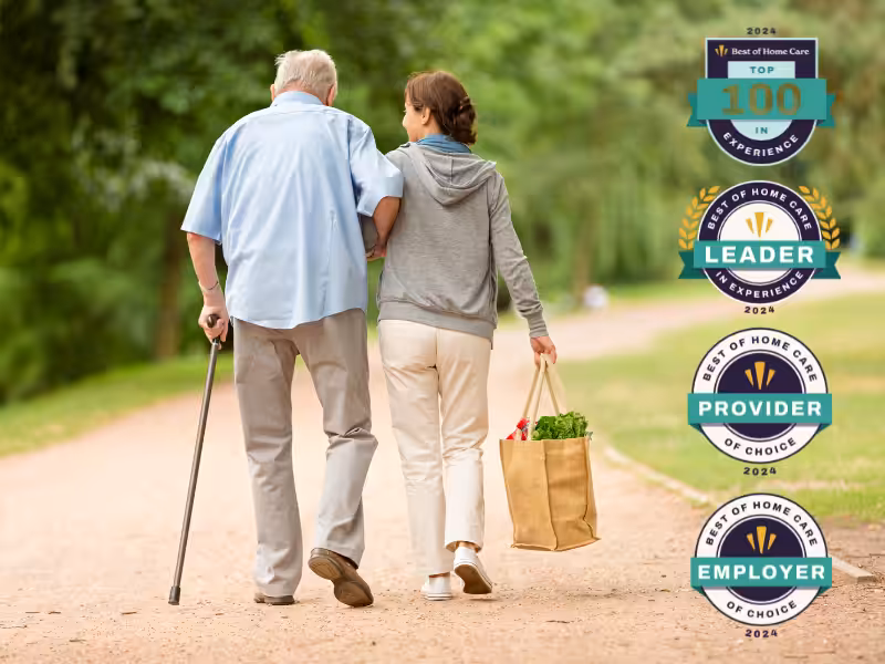 An elderly man with a cane walks arm-in-arm with a woman carrying groceries on a path. Four award badges for home care achievements in 2024 are displayed on the right side of the image.