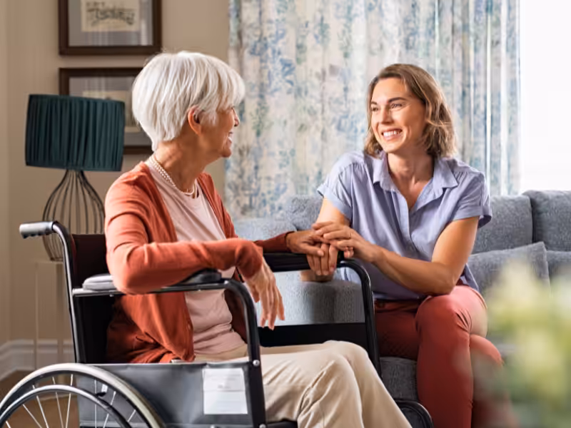 A smiling young woman sits on a couch holding hands with an older woman in a wheelchair, both engaged in a warm, cheerful conversation in a cozy living room.