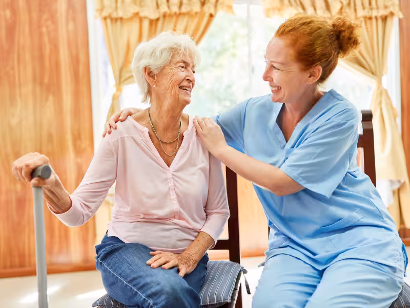 An elderly woman with a cane smiles while sitting next to a caregiver in blue scrubs. They share a warm, joyful moment together in a bright, home-like setting.