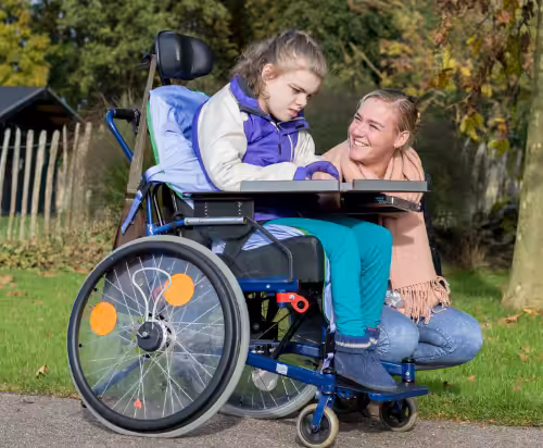 A young girl in a wheelchair sits outdoors, wearing a purple jacket and teal pants. A woman kneels beside her, smiling warmly. Trees and a fence are visible in the background on a sunny day.