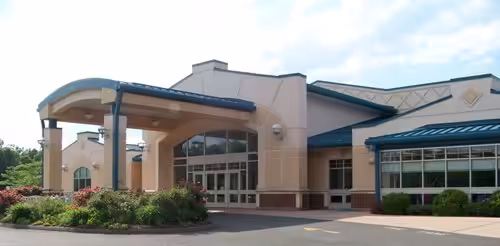 Single-story beige building with large windows, a covered entrance supported by columns, landscaped bushes, and flowering plants in front. The roof has teal accents and the sky is partly cloudy.