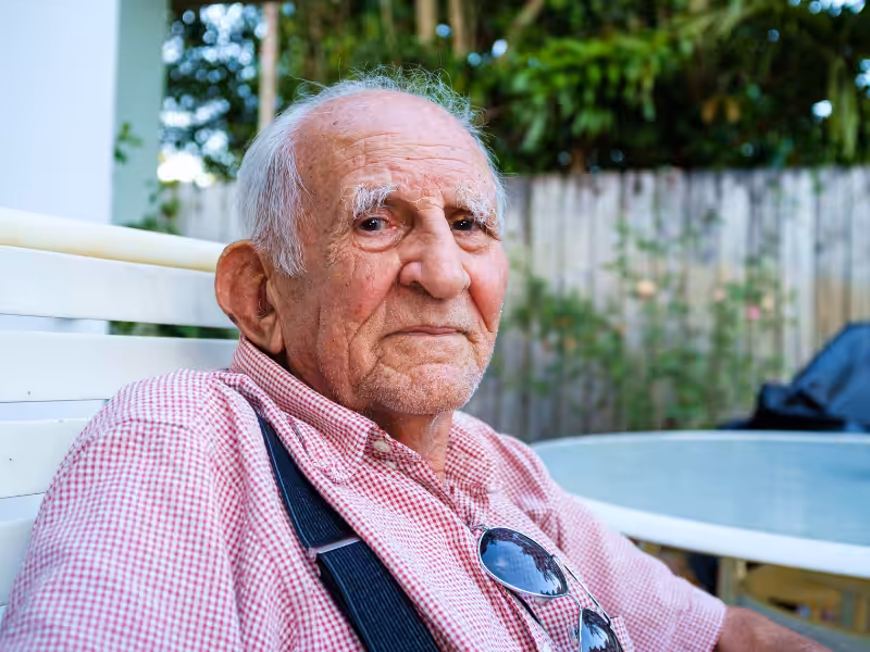 An elderly man with white hair and a pink checkered shirt sits outdoors on a white chair, looking at the camera. He wears suspenders and eyeglasses hang from his shirt. Green plants and a wooden fence are in the background.
