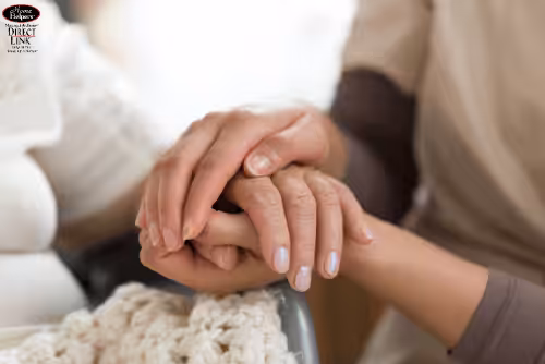 A close-up of one person gently holding the hand of another, who is covered with a knitted blanket, suggesting comfort, support, and care.