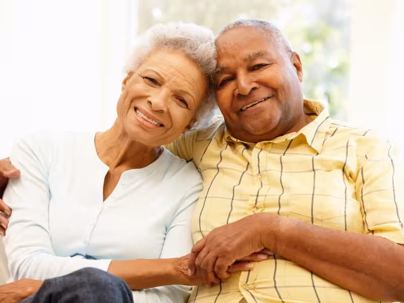 An elderly couple sits closely together on a couch, smiling warmly at the camera. The woman has gray hair and wears a light blue top, while the man wears a yellow plaid shirt. They appear happy and content.