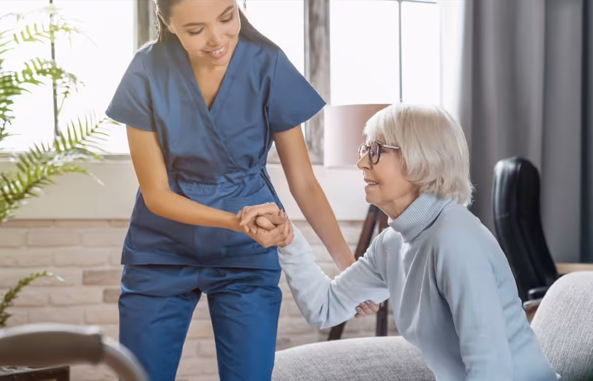 A caregiver in blue scrubs helps an elderly woman with white hair and glasses stand up from a couch in a well-lit living room.