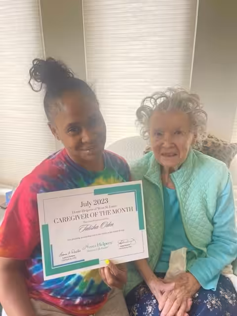 A woman in a tie-dye shirt holds a Caregiver of the Month certificate from July 2023, sitting beside an elderly woman in a mint green jacket. They are both smiling and seated indoors.