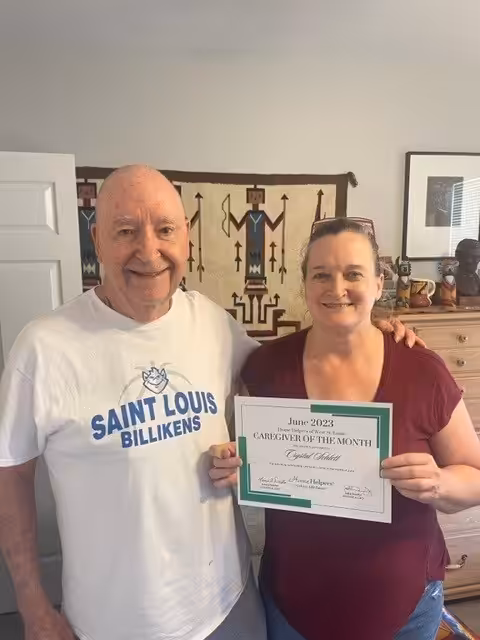 A smiling woman holds a “Caregiver of the Month” certificate while standing next to an older man in a Saint Louis Billikens shirt. They are indoors with a tapestry and various items in the background.