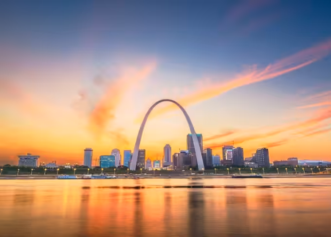 The Gateway Arch in St. Louis, Missouri, stands tall against a vibrant sunset sky, with city buildings and their reflections visible on the calm water in the foreground.