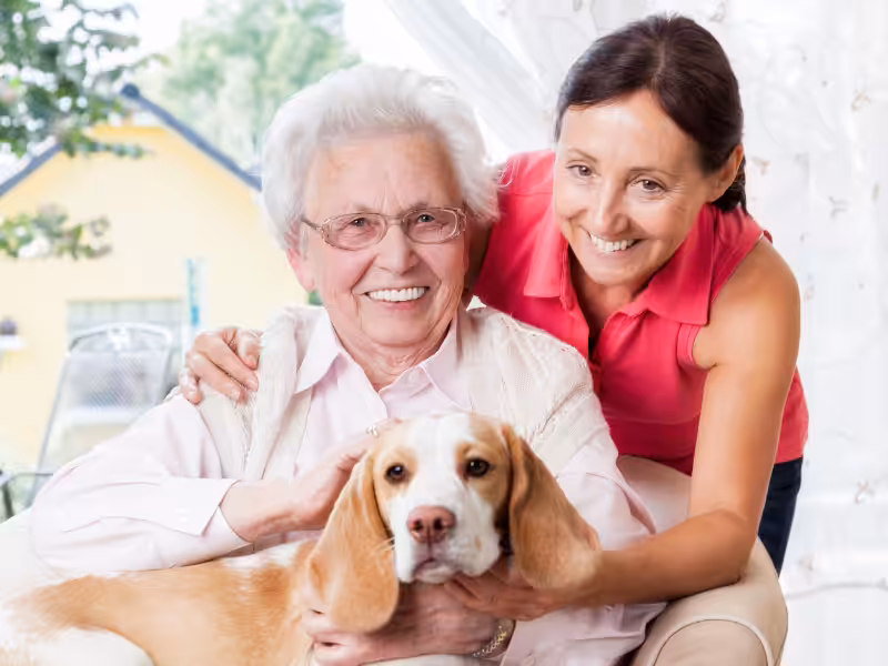 An elderly woman and a younger woman smile while sitting together indoors, with a beagle dog resting its head on the elderly womans lap. All three appear happy and relaxed.