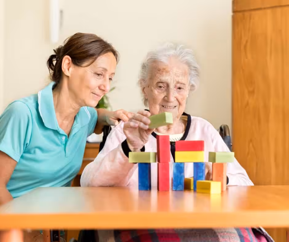 A younger woman sits beside an elderly woman who is smiling and stacking colorful wooden blocks on a table, both appearing engaged and happy in a cozy indoor setting.