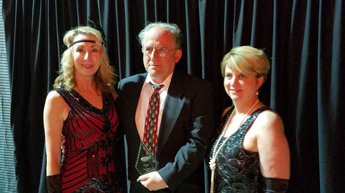 Three adults dressed in formal 1920s-style attire stand together in front of a black curtain. The man in the center holds a glass award and is flanked by two women wearing decorative dresses and accessories.