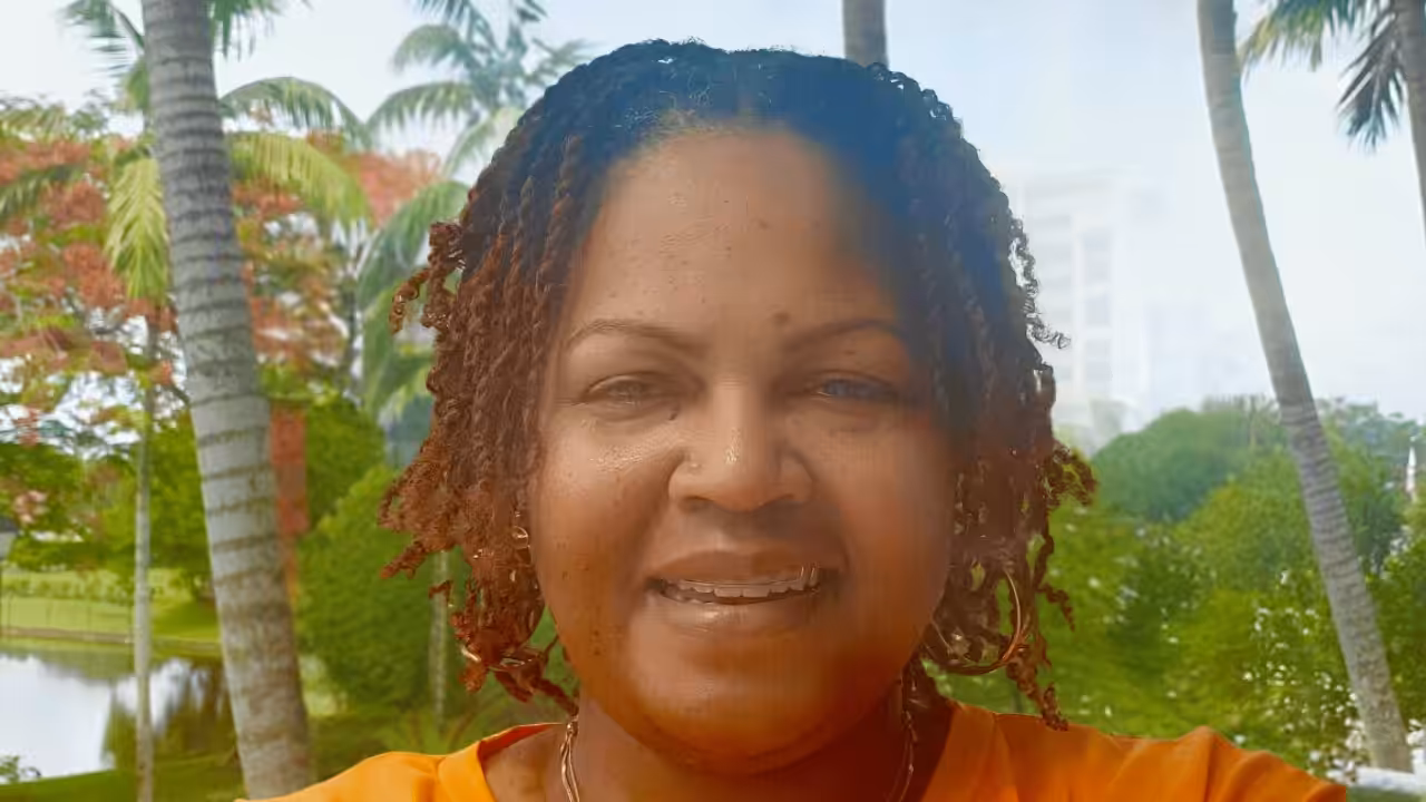 A woman with short, curly hair smiles outdoors, with palm trees, green foliage, and a pond in the background. She is wearing an orange shirt and a necklace, and the scene appears bright and sunny.