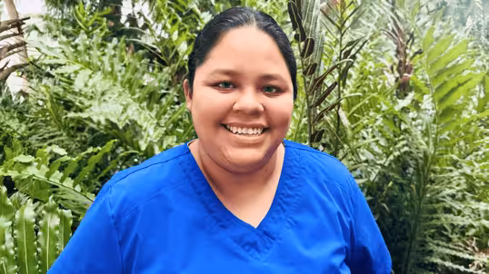 A person with dark hair pulled back, wearing a bright blue scrub top, smiles while standing in front of lush green plants outdoors.