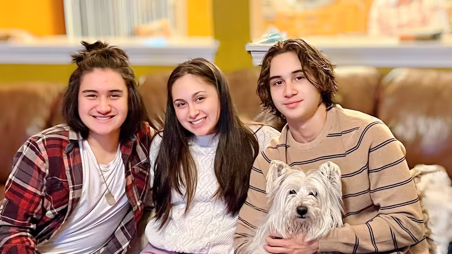 Three smiling young people sit close together on a couch, with the person on the right holding a small white dog. The background has warm yellow tones and a blurred interior setting.