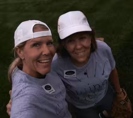 Two women wearing white caps and light purple shirts smile at the camera. They are outdoors on grass, and one woman is holding a baseball glove. Both have name tags on their shirts.