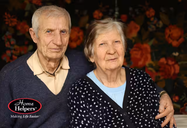 An elderly man and woman sit closely together, smiling warmly. The man wears a light shirt and beige sweater, while the woman wears a polka-dot sweater. The background features a floral pattern. Home Helpers logo is in the corner.
