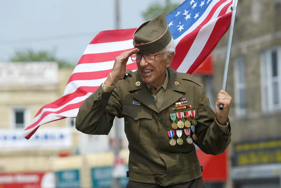 An elderly person in a decorated military uniform salutes while holding a U.S. flag over their shoulder, smiling in an outdoor setting with blurred buildings in the background.