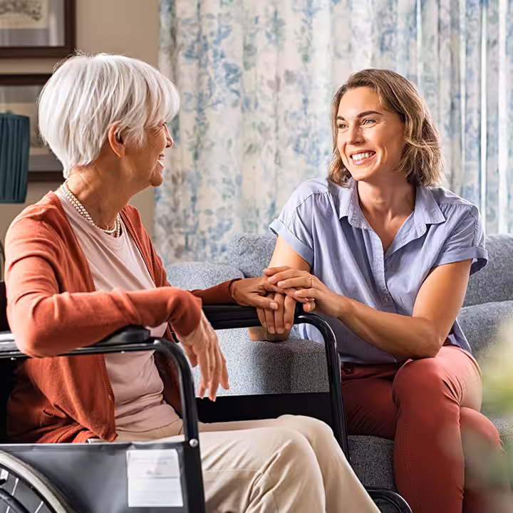 A smiling younger woman sits beside an older woman in a wheelchair, holding her hand. They are indoors, engaged in a warm, friendly conversation in a cozy, well-lit living room.