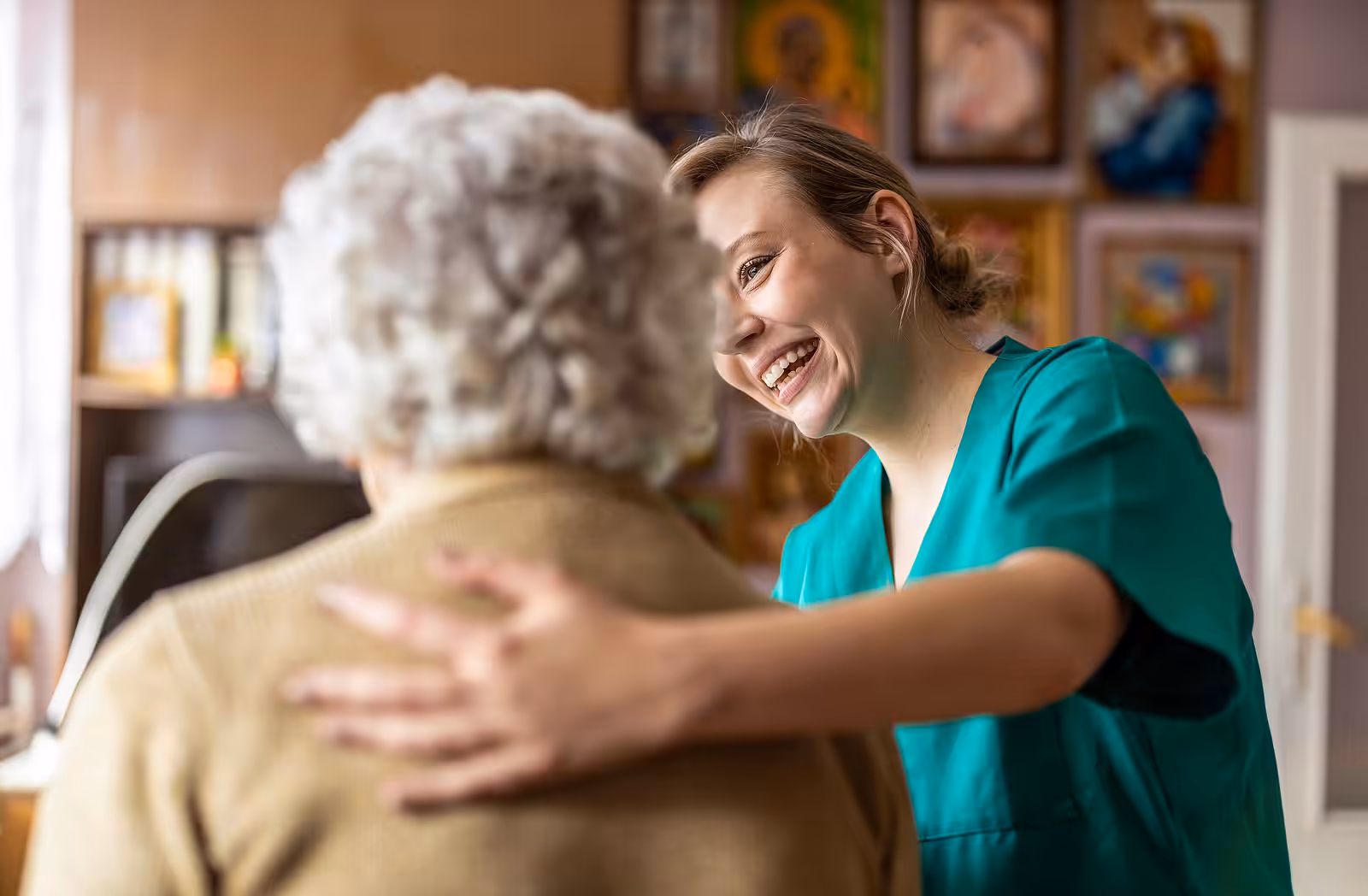 A smiling caregiver in green scrubs gently places a comforting hand on the shoulder of an elderly woman with gray hair, sitting together in a warmly decorated room with framed pictures on the wall.