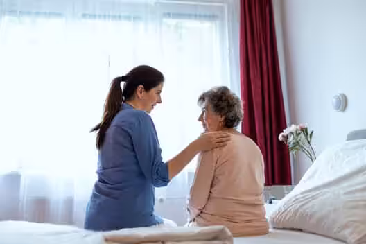 A young woman sits beside an older woman on a bed, gently touching her shoulder. They are facing each other and appear to be having a caring conversation in a bright, cozy room with flowers on a bedside table.