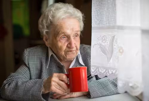 An elderly woman with white hair sits by a window, holding a red mug and gazing outside thoughtfully. She is wearing a checkered jacket and is partially illuminated by natural light coming through lace curtains.