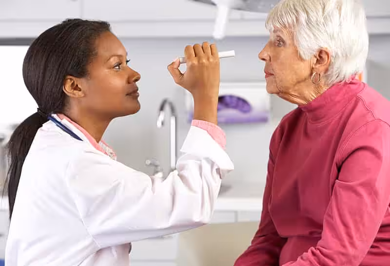 A doctor uses a penlight to examine the eyes of an older woman with short white hair, who is sitting on an exam table in a medical office.