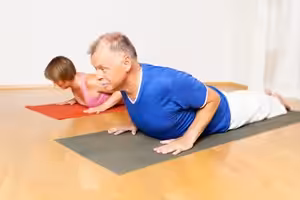 Two people practice yoga indoors on mats, performing the cobra pose. The person in front wears a blue shirt and white pants, while the person behind wears a pink top. Both appear focused and are in a bright room.