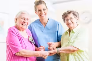 Three women, two elderly and one younger in a nurse uniform, stand close together indoors, smiling warmly and holding hands, suggesting a friendly and supportive environment.
