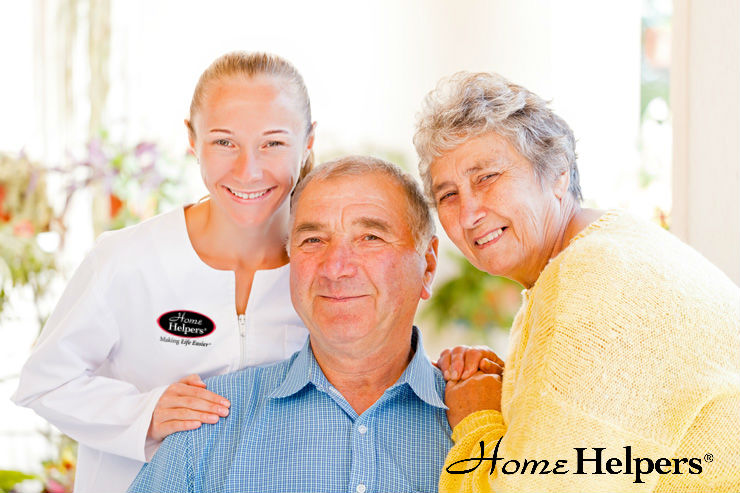 A smiling caregiver in a white uniform stands beside a happy elderly couple. The man sits while the woman stands with her hand on his shoulder. The Home Helpers® logo appears in the corner.
