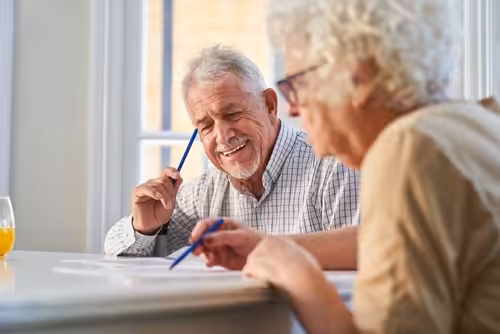 Two older adults sit at a table by a window, smiling and holding pens while working on papers together. A glass of orange juice is on the table beside them.
