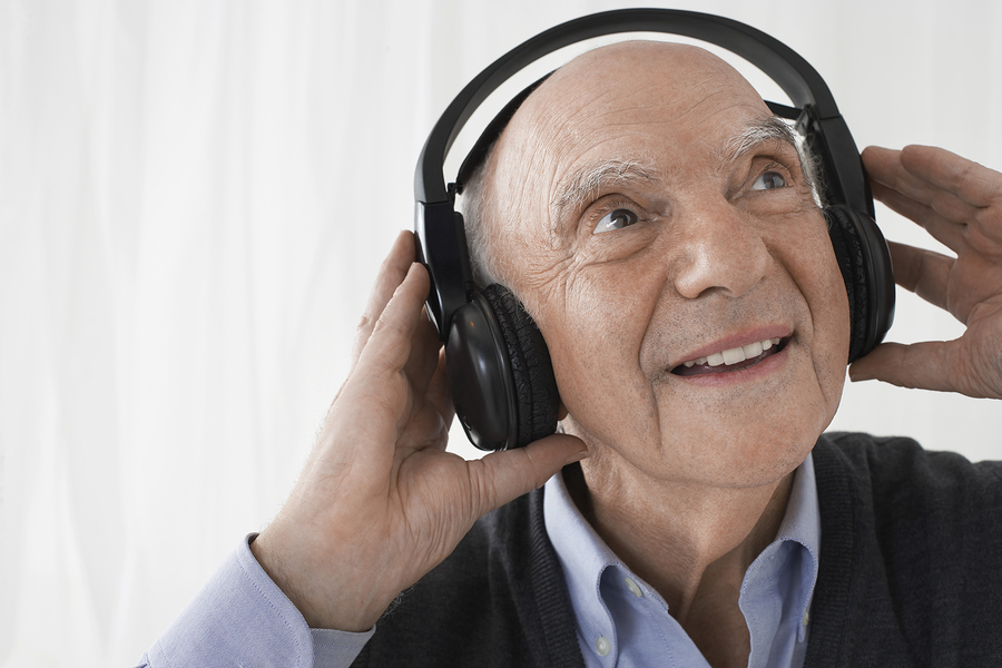 Smiling elderly man wearing large black headphones, holding them with both hands, and looking upward while enjoying music. He is dressed in a light blue shirt and dark sweater, against a light background.