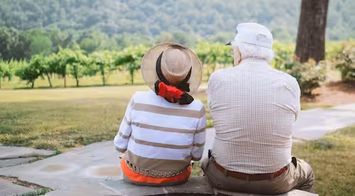 An elderly couple sits side by side on a stone path, facing a green vineyard and distant hills. They are wearing hats and casual clothes, enjoying a peaceful outdoor moment together.
