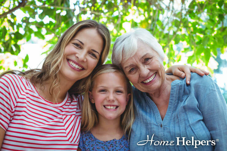 Three generations of women—an older woman, an adult woman, and a young girl—smiling together outdoors under leafy trees. The adult and older woman embrace the girl. Home Helpers logo appears in the bottom right corner.