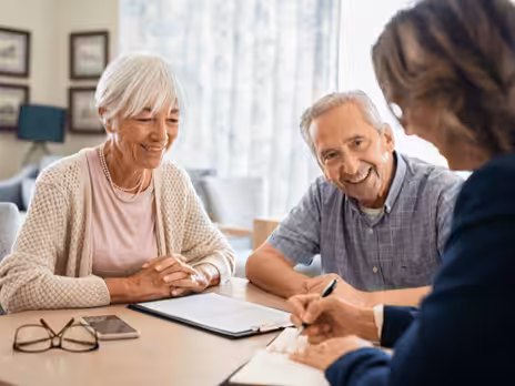 An elderly couple sits at a table, smiling and talking with a professional woman who is writing on a notepad. A smartphone and a clipboard are on the table in front of them.