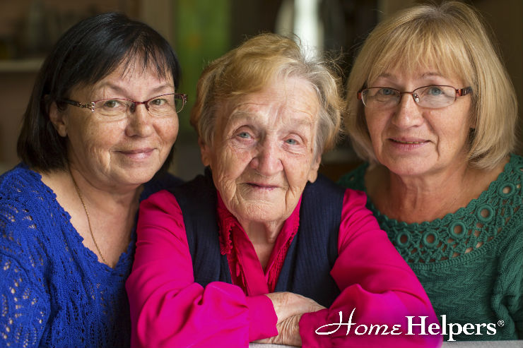 Three older women are sitting closely together, smiling warmly at the camera. The words Home Helpers appear in the bottom right corner.