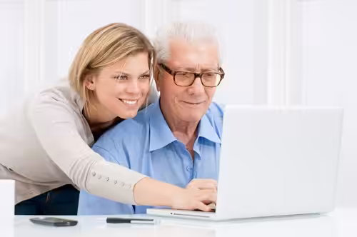 A smiling younger woman stands beside an older man with gray hair and glasses as they both look at a laptop screen. The woman points at the keyboard, helping the man use the computer.