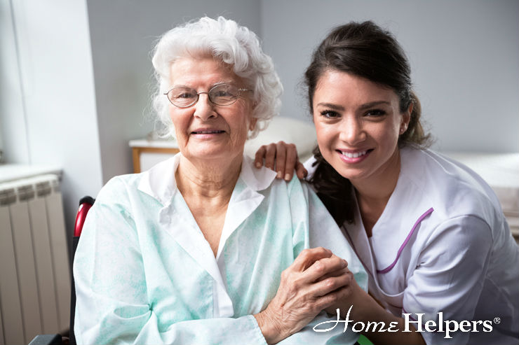 A smiling elderly woman in glasses sits beside a cheerful female caregiver in a white uniform. They are indoors, holding hands, with the Home Helpers logo in the corner.