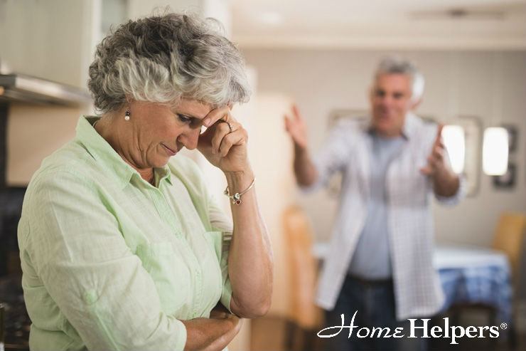 An older woman looks upset with her hand on her forehead while an older man gestures and speaks to her in the background. The image has the Home Helpers logo in the bottom right corner.