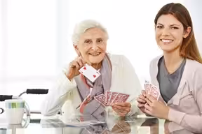 An elderly woman and a young woman are sitting at a table, smiling and holding playing cards. The elderly woman is showing an ace of hearts. A walker is visible in the background.