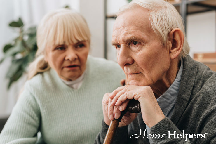 An elderly man with a cane sits thoughtfully, while an elderly woman sits beside him, looking concerned. Both are indoors, and the Home Helpers logo is visible in the bottom right corner.