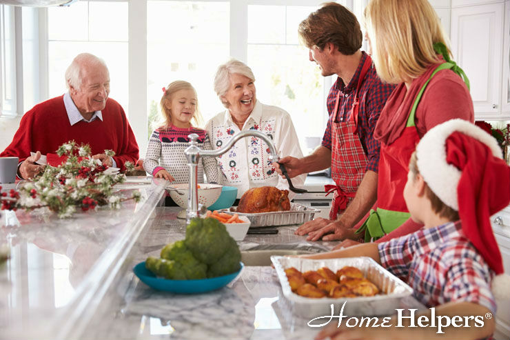 A multigenerational family prepares and smiles around a kitchen island with holiday food. An older couple, two children, and two adults wear festive clothes. The counter is decorated with seasonal greenery.
