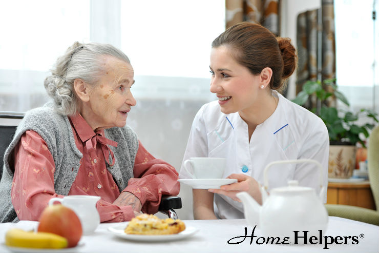 A smiling caregiver in uniform offers a cup of tea to an elderly woman sitting at a table with fruit and pastries. Both women are engaged and happy. The Home Helpers® logo is visible in the corner.