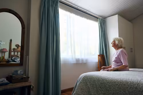 An elderly woman sits on the edge of a bed in a softly lit bedroom, gazing thoughtfully out a window with light blue curtains. A mirror and dresser are visible on the left side of the room.