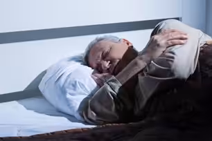 An older man with gray hair sleeps on his side in bed, covered with a blanket and resting his head on a white pillow.