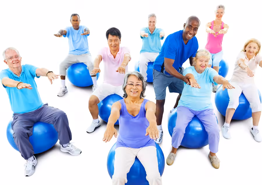 A group of older adults, both men and women, sit on exercise balls with their arms extended, participating in a fitness class led by an instructor, all smiling and wearing colorful workout clothes.