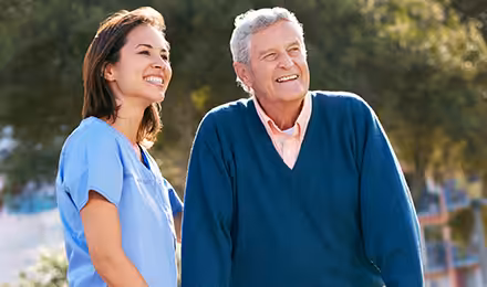 A smiling healthcare worker in blue scrubs stands outdoors next to an older man in a blue sweater. They both look ahead happily, with trees and sunlight in the background.