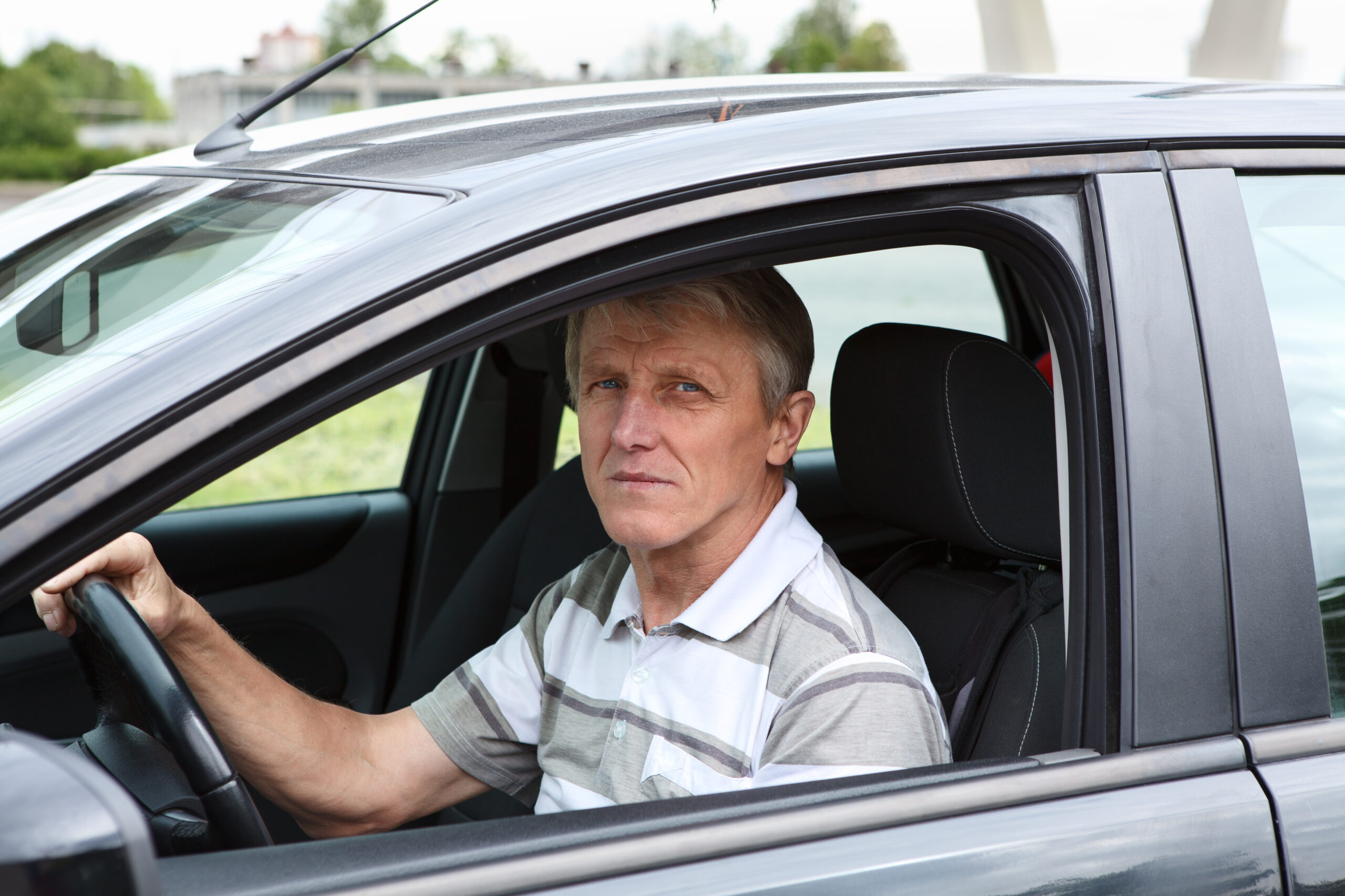 An older man with gray hair wearing a striped polo shirt sits in the drivers seat of a car, looking out of the open window with a serious expression.
