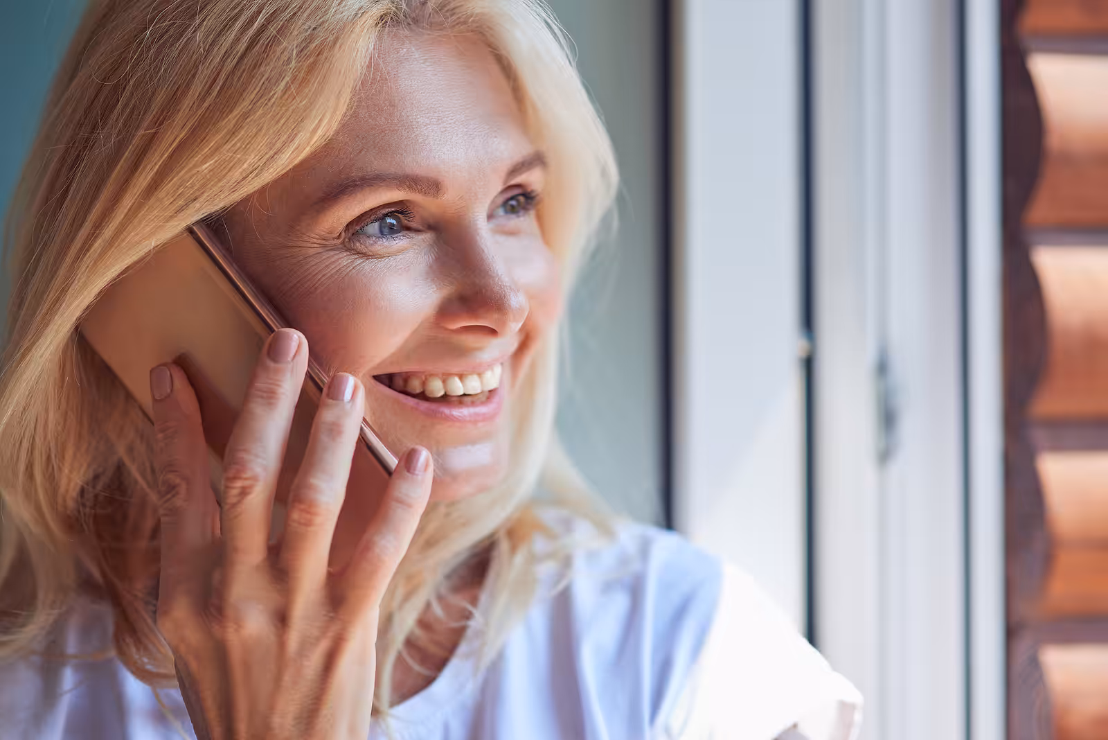 A smiling woman with blonde hair holds a smartphone to her ear while talking on the phone, standing by a window with sunlight on her face.