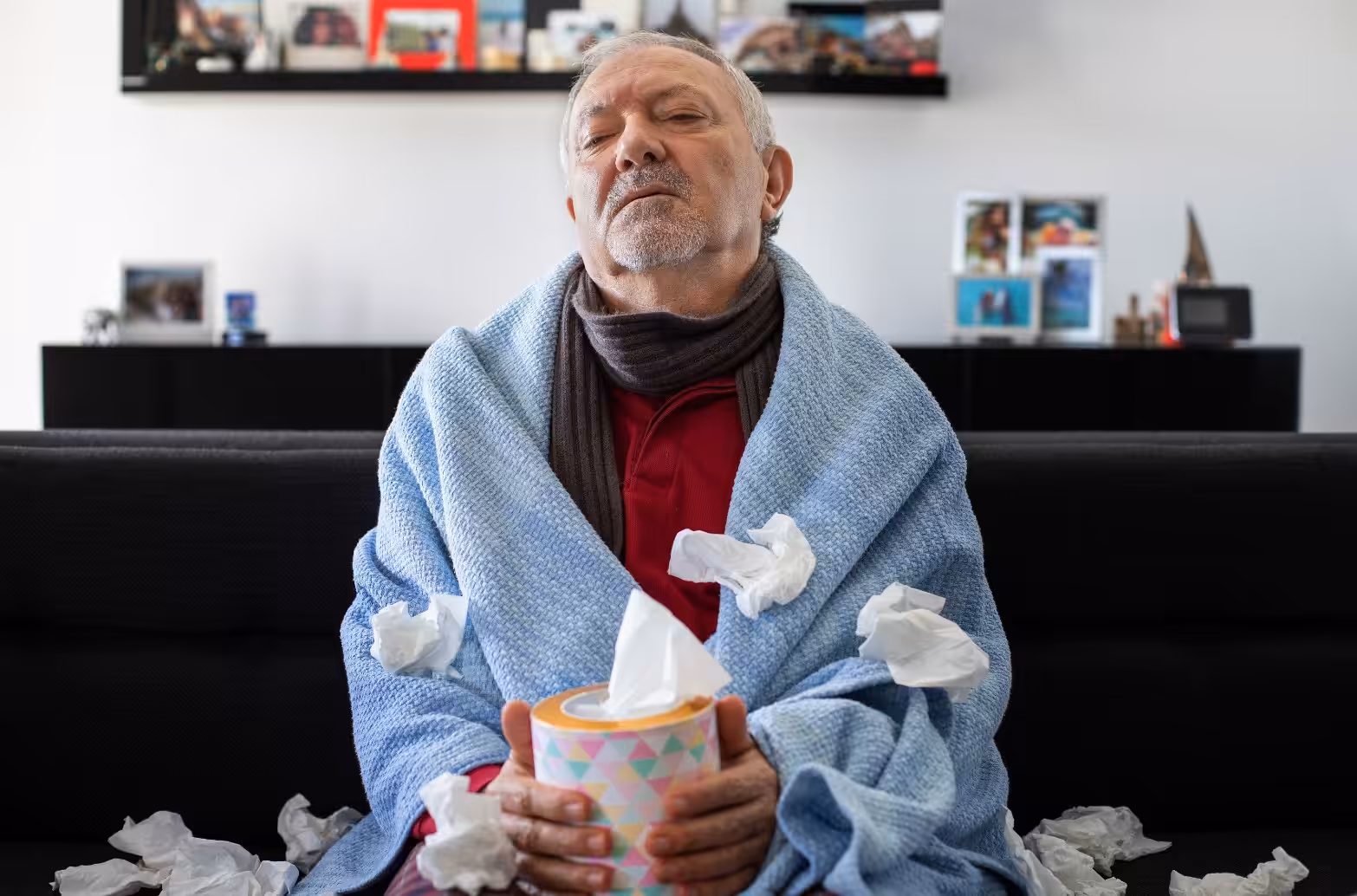 An older man sits on a couch wrapped in a blue blanket, wearing a scarf, and holding a box of tissues. Used tissues are scattered on his lap and the couch, suggesting he is feeling unwell.